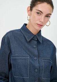 Camicia di jeans di colore blu scuro, con chiusura a bottoni frontale, colletto classico, due tasche sul petto e cuciture bianche in evidenza.