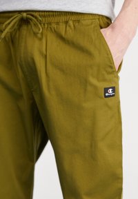 Olivgröna joggerbyxor i bomull med elastisk midja och dragsko. Har sidofickor och en liten logotyppatch på vänster sida.