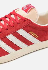 Röd mockaskor med vita läderstrimmor, texturerad gummisula och guld "Gazelle" broderi på sidan. Har röda skosnören och tunga.