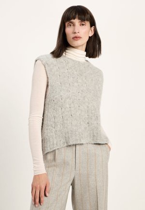 Woman wearing a light gray fuzzy sleeveless sweater over a white turtleneck and light gray pinstriped pants against a plain background.