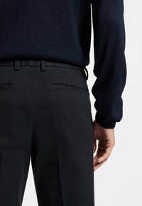 Czarne wizytowe spodnie o gładkiej fakturze, z plisami z przodu i dwiema tylnymi kieszeniami. Nosi się je z granatowym swetrem. Ręka oparta na boku.