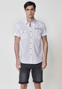 Camisa blanca de manga corta con dos bolsillos frontales, botones negros y detalles estampados. Combinada con pantalones cortos de mezclilla oscura con dobladillos enrollados.