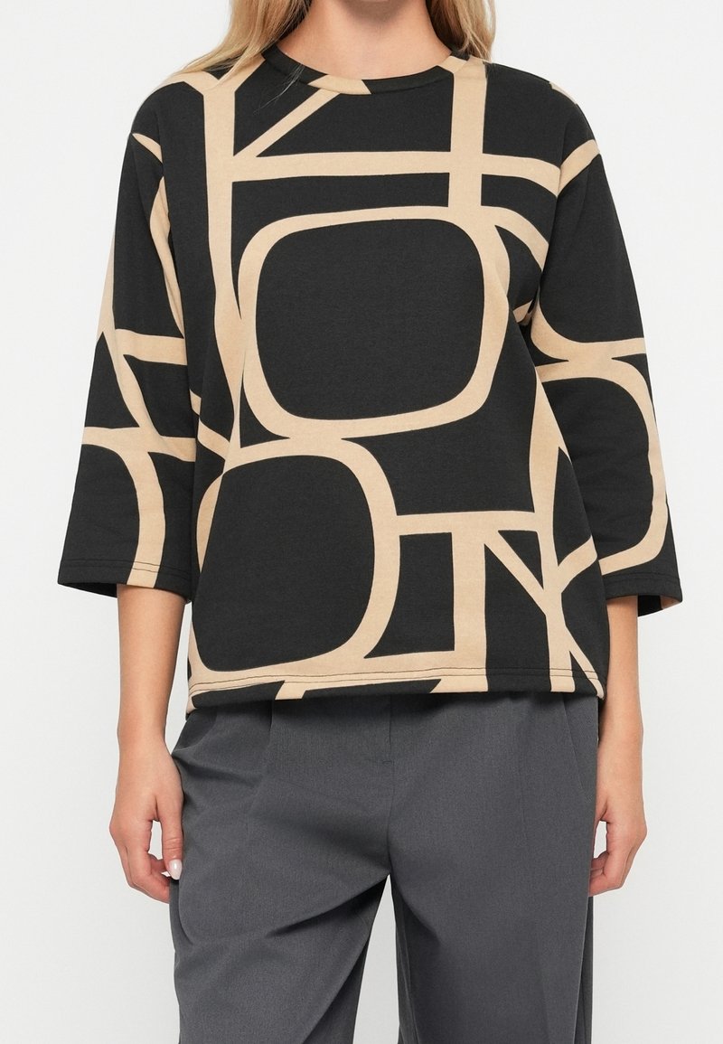 Femme portant un haut ample noir et beige à motifs avec des manches trois-quarts et un pantalon gris sur un fond uni.