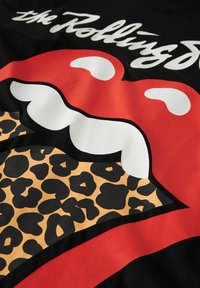 Crvene usne Rolling Stones s bijelim zubima i jezikom u leopard printu na crnoj tkanini s bijelim tekstom "the Rolling" iznad.