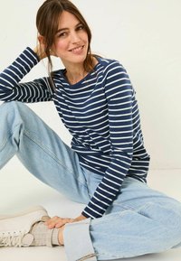 Young woman smiling, sitting cross-legged, wearing a navy and white striped long-sleeve shirt, light blue jeans, beige socks, and white sneakers.