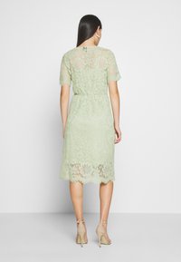 Robe en dentelle vert menthe à manches courtes, avec des motifs floraux et une longueur au genou. Le dos présente un détail en trou de serrure et une taille cintrée.