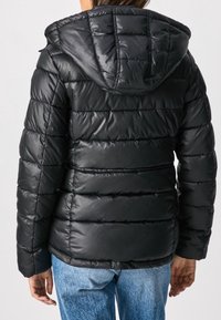 Svart puffjacka med huva, med en quiltad design och glänsande yta. Ärmarna är åtsittande och matchas med blå denimjeans.