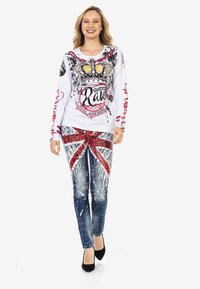 Langärmliges weißes Hemd mit Kronen-Grafik und roten Akzenten, kombiniert mit blauen Jeans, die ein rotes X-Muster und Farbspritzdetails aufweisen.