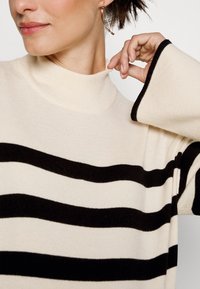 Krémový svetr s černými vodorovnými pruhy, vysokým límcem a širokými rukávy. Měkká textura s žebrovanými detaily na okrajích rukávů.