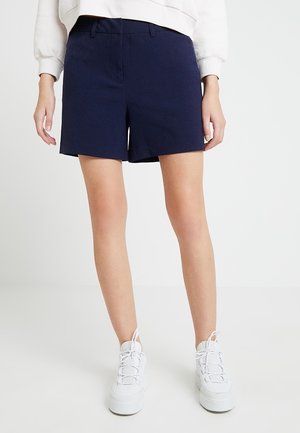 Kvinde iført marineblå skræddersyede shorts, hvid sweatshirt og hvide platformssneakers stående mod en ensartet lys baggrund.