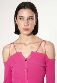 Cardigan aderente in maglia a coste di un vivace rosa, con design scivolato sulle spalle, chiusura con bottoni e spalline in perfetto accostamento metallico.