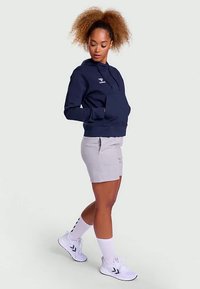 Jūros pajėgų spalvos gobtuvinė palaidinė su priekinėmis kišenėmis ir logotipu; derinama su šviesiai pilkomis šortais. Balti sportiniai batai ir kojinės užbaigia aprangą.