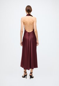 Robe bordeaux à col licou avec un dos décolleté, réalisée en satin lisse. Elle est dotée de liens réglables sur les côtés et d'une jupe fluide atteignant mi-mollet.