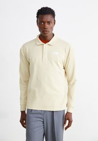 Lys gul langærmet polo-shirt med en afslappet pasform, der har en to-knaps stolpe og et hvidt logo på venstre bryst. Grå bukser.