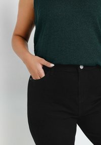 Jeans neri con un design attillato e tasche laterali, abbinati a un top senza maniche di un verde scuro texture. Dettaglio sottile in bottone argentato visibile.