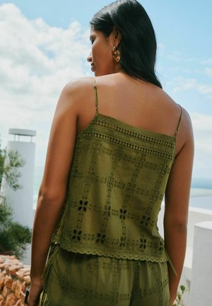 Woman with long dark hair wearing gold hoop earrings and a green embroidered sleeveless top outdoors against a blue sky background.
