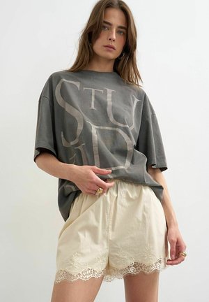 Kvinde iført oversized grå t-shirt med store bogstaver og cremefarvede shorts med blonder, stående mod en ensfarvet hvid baggrund.