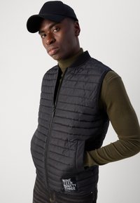 Zwarte gewatteerde mouwloze vest met een ritsluiting, horizontale stiksels en zijzakken. Draagt over een groene coltrui.
