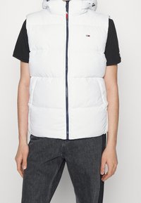 Vit pufferväst med hög krage och dragkedjestängning, som har en marinblå accent längs dragkedjan och en liten logotyp på bröstet.