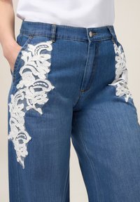 Pantalones de mezclilla con acentos florales bordados en blanco en los bolsillos delanteros, con un corte de cintura alta y piernas rectas.