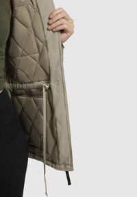 Lichte, beige gewatteerde jas met trekkoord in de taille, drukknoopsluiting en een gladde textuur. Voering zichtbaar, met ontwerpdetails.