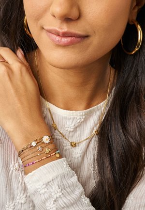 Frau mit goldenen Creolen, geschichteten goldenen Halsketten und mehreren zarten Armbändern mit Blumen- und Anhängerdesigns am Handgelenk.