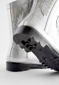 Metallisch silberne kniehohe Stiefel mit einer schwarzen Gummisohle und strukturiertem Profil; mit einem Flammenmuster an der Außenseite.