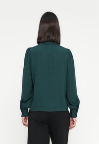 Mørkegrønn bluse med lange ermer, puffede skuldre, teksturert stoff og krage. Bakken viser en rett søm og løs passform.