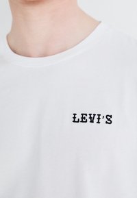 Weißes Baumwoll-T-Shirt mit einem schwarzen gestickten "LEVI'S"-Logo auf der linken Brustseite. Klassisches Rundhalsausschnitt-Design, glatte Textur.