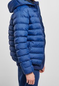 Blå puffjacka med en glänsande yta, med huva, horisontella quiltade sektioner och framfyllning. Sidofickor ingår.