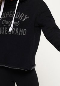 Svart kortärmad hoodie med vita dragsnören. Har metallisk text "SUPERDRY Original True Brand" framtill. Mjuk textur.