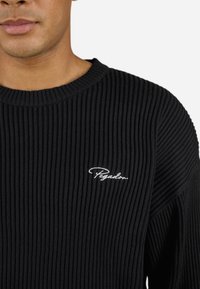 Sort ribbet sweater med rund halsudskæring. Har broderet hvid branding på venstre bryst. Teksturen virker blød og struktureret.