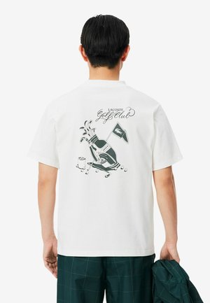 Homme portant un t-shirt blanc avec un graphisme de sac de golf, clubs, balles et un drapeau Lacoste ainsi que le texte "Lacoste Golf Club" dans le dos.