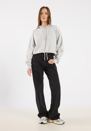 Felpa grigia chiara con cappuccio e tasca frontale, abbinata a pantaloni neri a gamba larga e sneakers multicolori su uno sfondo neutro.