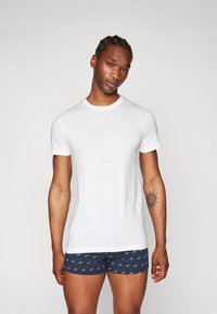 T-shirt en coton blanc à manches courtes, de forme fitted. Porté avec un short boxer à motifs bleu marine, mettant en avant un logo blanc répété.