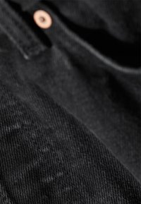 Gros plan d’un tissu en denim noir montrant une texture détaillée, des coutures et un petit rivet en cuivre.