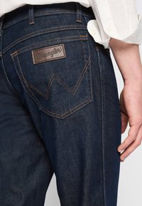 Wrangler mörka denimjeans har en läderlogotyp, kontrasterande sömmar och en distinkt bakfickdesign med ett zigzag-mönster.