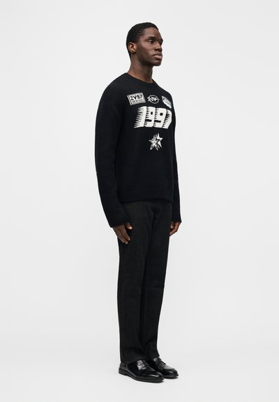 Černý svetr s grafickými motivy včetně nápisu „1997“, nošený přes tmavé kalhoty a černé boty. Svetr má texturovaný povrch a žebrované manžety.