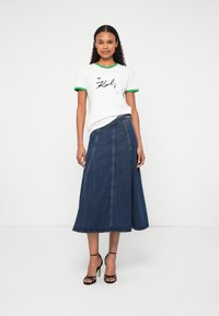 Bijela majica s detaljima u zelenoj boji na izrezu koji sadrži crni grafički tekst. Duga traper suknja tamnoplave boje. Model nosi crne sandale s potpeticama.