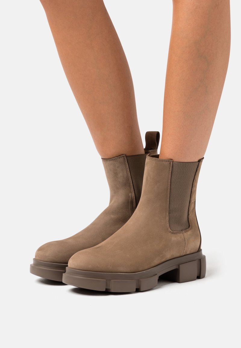 Bottes en daim taupe montant à la cheville avec semelles épaisses en caoutchouc marron, portées sur jambes nues sur fond blanc.