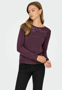 Un pullover en tricot violet à manches longues, orné de sequins le long de l'encolure. Associé à un pantalon noir, texture lisse.