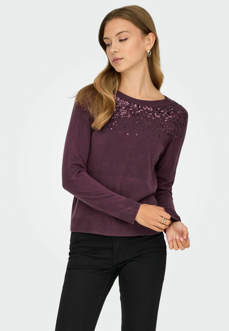 Un pullover en tricot violet à manches longues, orné de sequins le long de l'encolure. Associé à un pantalon noir, texture lisse.