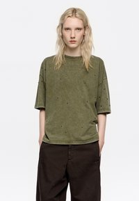 Camiseta de manga corta verde oliva con un corte relajado, que presenta adornos negros dispersos y una tela suave y texturizada.