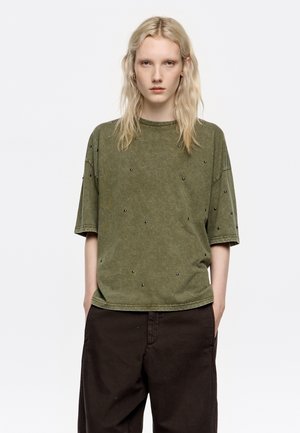 Camiseta de manga corta verde oliva con un corte relajado, que presenta adornos negros dispersos y una tela suave y texturizada.