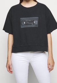 Mustaväri oversized lyhythihainen collegepaita, jossa on graafinen printti VHS-nauhasta. Raaka viimeistely, pehmeä kangas ja rento istuvuus. Yhdistetty valkoisiin farkkuihin.