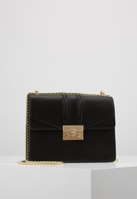 Borsa a tracolla in pelle nera lavorata con chiusura rettangolare dorata e tracolla coordinata in oro, posizionata su una superficie bianca contro uno sfondo grigio.