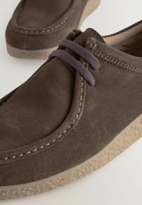 Brune suedesko med rund tå, syninger og grå snørebånd. Sålen er struktureret med en lys beige farve.