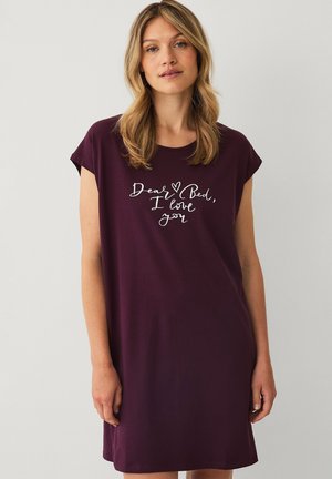 Vrouw met blond haar die een donkerpaarse jurk met korte mouwen draagt met witte tekst ""Dear Bed, I love you"" aan de voorkant.