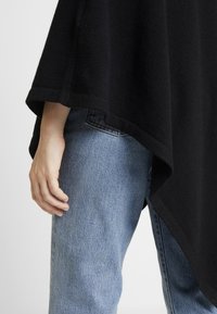 Schwarzer, gestrickter Poncho liegt über einer Hand, die auf blauen Jeans aus Denim ruht. Der Stoff hat eine weiche Textur und eine breite, fließende Form.