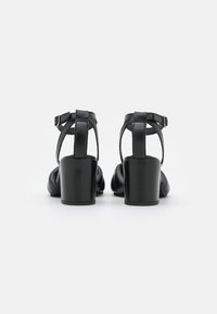 Sandales en cuir noir à talon carré avec un dos ouvert et une bride ajustable à la cheville, présentant une texture lisse et un design minimaliste.